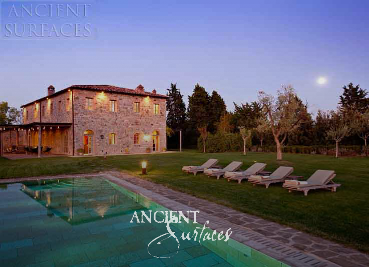 Ancient Surfaces Arcane Stone pool coping and terrace paving at dusk beside a lit stone farmhouse